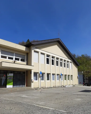 Ein modernes, zweigeschossiges Gebäude mit einer hellen Fassade und großen Fenstern. Vor dem Gebäude befindet sich ein Parkplatz mit mehreren Parkplätzen. Der Himmel ist klar und blau.