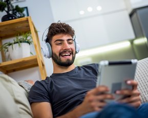 Junger Mann mit Kopfhörern, der auf einem Tablet sitzt und lächelt, während er entspannt auf einem Sofa ist.