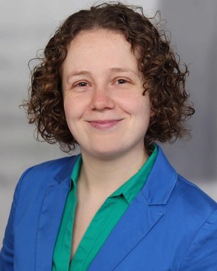 Eine Person mit lockigem Haar trägt einen blauen Blazer über einem grünen Hemd. Sie lächelt freundlich in die Kamera.