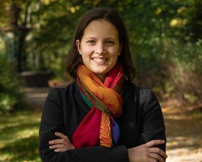 Eine lächelnde Frau mit einem bunten Schal steht mit verschränkten Armen in einem herbstlichen Park.