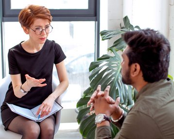Klinische Psychologie Fernstudium | SRH Fernhochschule Eine Frau mit Brille sitzt auf einem Stuhl und spricht mit einem Mann, der ihr gegenüber sitzt. Beide scheinen in ein Gespräch vertieft zu sein, umgeben von Pflanzen und natürlichem Licht.