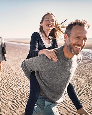 Eine Familie spaziert am Strand. Ein Mann trägt ein Mädchen auf dem Rücken, während zwei Frauen und ein Junge in der Nähe gehen. Alle lächeln und genießen den Tag.