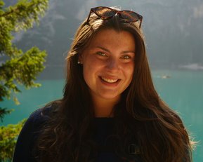 Eine Frau mit langen, braunen Haaren und Sonnenbrille lächelt vor einem klaren, türkisfarbenen See und bewaldeten Bergen.