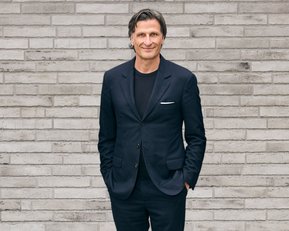 Mann in einem dunklen Anzug steht mit verschränkten Händen vor einer grauen Ziegelwand.