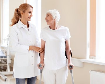 Rehabilitationspsychologie Studium | SRH Fernhochschule Eine Klinikmitarbeiterin unterstützt eine ältere Patientin, die mit Gehhilfen steht. Beide lächeln sich an, während sie in einem hellen Raum stehen.