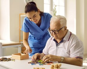 Eine Pflegekraft hilft einem älteren Mann beim Puzzeln am Tisch, während er konzentriert an den Teilen arbeitet.