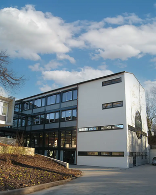 Moderne Architektur eines mehrstöckigen Gebäudes mit großen Fenstern und einem weißen Fassadenabschnitt, umgeben von Bäumen und einem klaren Himmel.