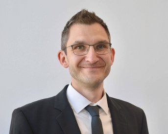 Ein Mann in einem schwarzen Anzug mit Krawatte und Brille lächelt freundlich. Der Hintergrund ist neutral und hell.