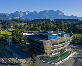Moderne Architektur eines Gebäudes mit geschwungener Fassade, umgeben von grünen Wiesen und Bäumen, im Hintergrund eine Berglandschaft mit hohen Gipfeln und klarem Himmel.