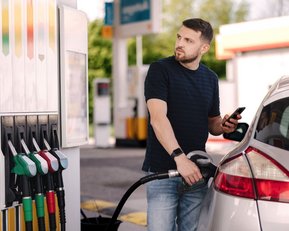 © Aleksandr – stock.adobe.com Ein Mann tankt sein Auto an einer Tankstelle und schaut auf sein Smartphone. Mehrere Zapfsäulen sind im Hintergrund sichtbar.