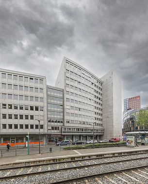 Moderne Bürogebäude mit Glasfassade und asymmetrischer Architektur, umgeben von Straßen und Schienen. Der Himmel ist bewölkt, was eine gedämpfte Lichtstimmung erzeugt.
