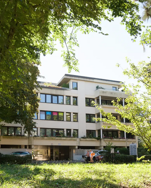 Mehrstöckiges Gebäude mit großen Fenstern und Balkonen, umgeben von Bäumen und Grünflächen. Im Vordergrund sind zwei Personen in orangefarbener Kleidung zu sehen.