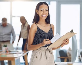 Eine Frau mit Clipboard und Notizen lächelt in einem modernen Büro, während Kollegen im Hintergrund an einem Tisch arbeiten.