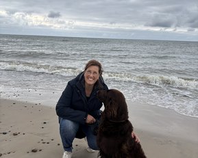 Eine Frau kniet am Strand und lächelt, während sie ihren braunen Hund streichelt. Im Hintergrund sind Wellen und bewölkter Himmel zu sehen.