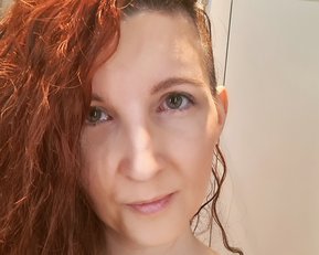 Eine Person mit langen, lockigen, roten Haaren schaut direkt in die Kamera. Sie trägt ein schwarzes Oberteil und hat einen neutralen Gesichtsausdruck.