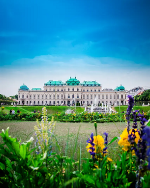 Das Schloss Belvedere mit grünen Dächern und barocker Architektur, umgeben von einem gepflegten Garten mit bunten Blumen und Springbrunnen. Der Himmel ist klar und blau.