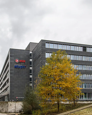 Moderne Bürogebäude mit grauer Fassade und großen Fenstern, beschriftet mit "RSO" und "alupr". Ein gelber Baum steht im Vordergrund, umgeben von grüner Vegetation.