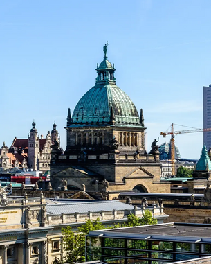 Die Kuppel eines historischen Gebäudes mit grünem Dach und Statue auf der Spitze, umgeben von weiteren architektonischen Strukturen und einem klaren blauen Himmel.