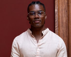 Ein junger Mann mit kurzen, geflochtenen Haaren und Brille trägt ein helles Hemd und steht vor einer roten Wand.