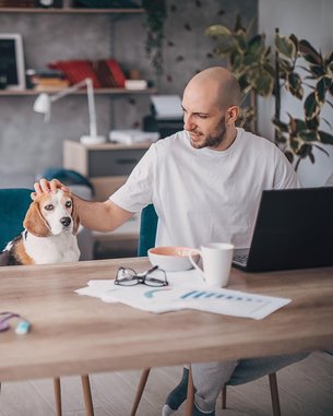 Ein Mann mit Glatze streichelt einen Beagle, während er an einem Tisch mit einem Laptop und einer Tasse Kaffee arbeitet. Auf dem Tisch liegen auch Unterlagen und eine Brille.