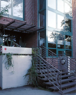 Eingang zur Caritas Akademie mit einer Treppe und einem modernen, verglasten Gebäude. Eine Pflanzenranke ist an der Wand sichtbar.