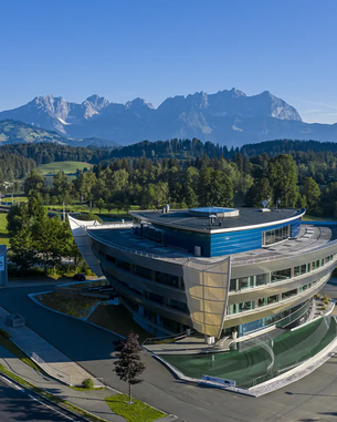 Moderne Architektur eines Gebäudes mit geschwungener Fassade, umgeben von Bäumen und Wiesen, im Hintergrund sind majestätische Berge zu sehen. Der Himmel ist klar und blau.