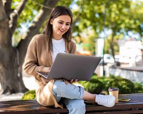 Eine junge Frau sitzt auf einer Bank im Freien und arbeitet an einem Laptop, während sie einen Kaffeebecher neben sich hat.