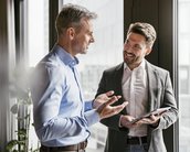 Steuerlastveränderung durch den neuen Koalitionsvertrag Zwei Männer in formeller Kleidung stehen in einem modernen Büro und führen ein Gespräch. Einer hält ein Tablet, während der andere gestikuliert. Große Fenster im Hintergrund lassen viel Tageslicht herein.
