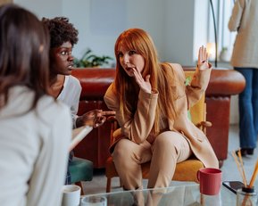 Drei Frauen sitzen in einem Café und unterhalten sich. Eine Frau mit langen roten Haaren gestikuliert lebhaft, während die anderen aufmerksam zuhören.