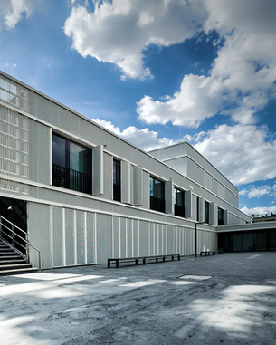 Moderne Architektur eines Gebäudes mit einer strukturierten Fassade, großen Fenstern und einem klaren, minimalistischen Design. Der Himmel ist teilweise bewölkt, was einen hellen Kontrast zur Bauweise bietet.