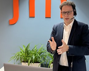 Ein Mann mit Headset steht vor einem Laptop und spricht, während im Hintergrund das Logo "srl" und eine Pflanze sichtbar sind.