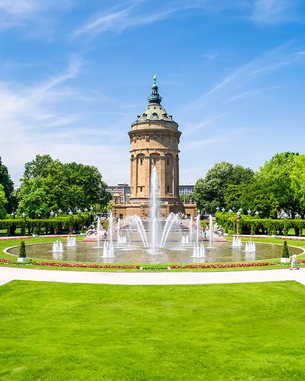 Der Wasserturm in Mannheim umgeben von einem gepflegten Park mit Brunnen und grünen Rasenflächen. Der Himmel ist blau mit wenigen Wolken.