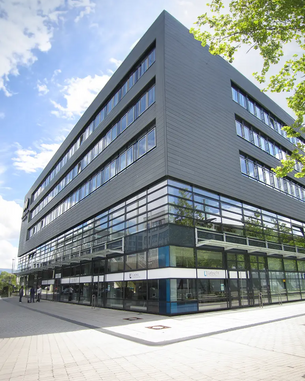 Modernes, mehrstöckiges Gebäude mit großen Glasfenstern und einem klaren, geometrischen Design. Die Fassade ist in dunklen Farben gehalten, umgeben von Bäumen und einem gepflasterten Gehweg.