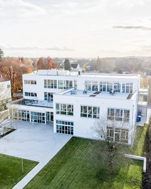 Moderne, mehrstöckige Gebäude mit großen Fenstern und einem begrünten Außenbereich. Die Architektur ist klar und funktional, umgeben von Bäumen und Rasenflächen.