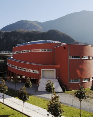 Moderne Architektur mit einem roten Rundbau und einem gläsernen Gebäude, umgeben von einer gepflegten Grünfläche und Bäumen, vor einer bergigen Kulisse.