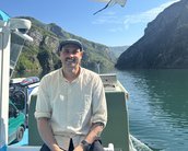 Ein Mann sitzt auf einem Boot und lächelt. Im Hintergrund sind grüne Berge und ein ruhiger Wasserweg zu sehen. Der Himmel ist klar und sonnig.